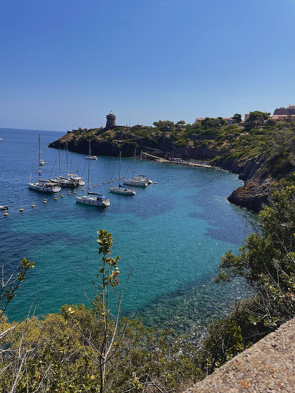 Barche a vela ormeggiate in una baia dall'acqua turchese, con una torre sul promontorio